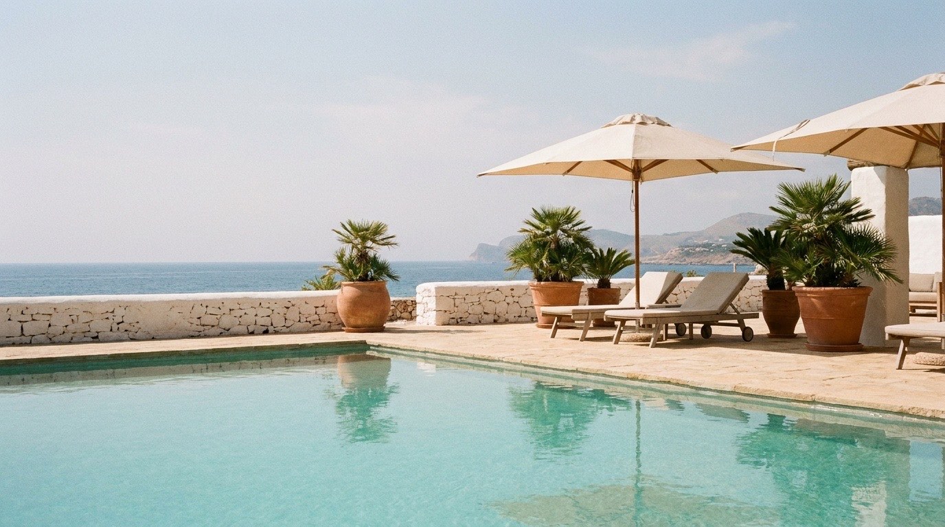 A still turquoise pool on a stone deck with linen umbrellas and the sea horizon beyond